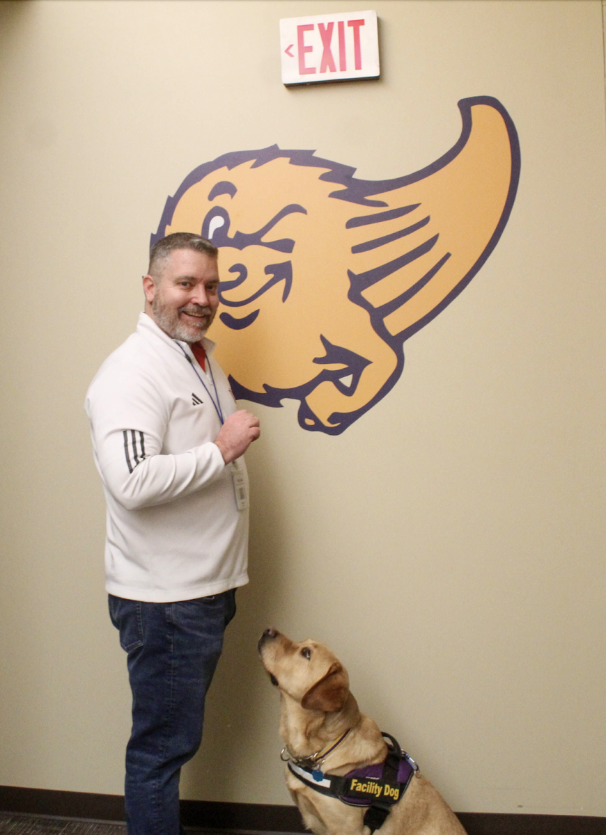 Blake Wegenke and Arlo posing infront of the Comets’ sign in student services. Wegenke is posing as if he is going to give Arlo a treat, and is helping him stay relaxed while the photo is being taken.